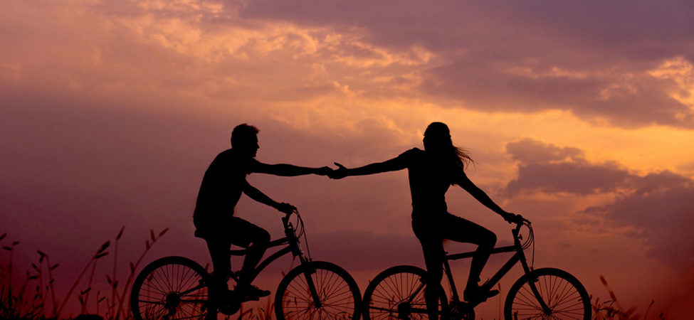 Mann og kvinne sykler sammen under solnedgangen og holder hender