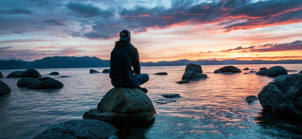 Em mann sitter på en stein i vannkanten og ser utover havet under en solnedgang