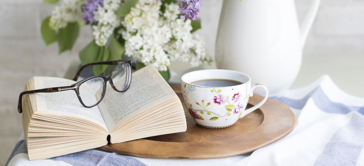 Bok oppslått på et bord med blomster og kaffekopp.