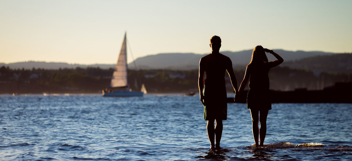 Silhuetten til to mennesker som står så vidt uti vannet i Oslofjorden og holder hender.