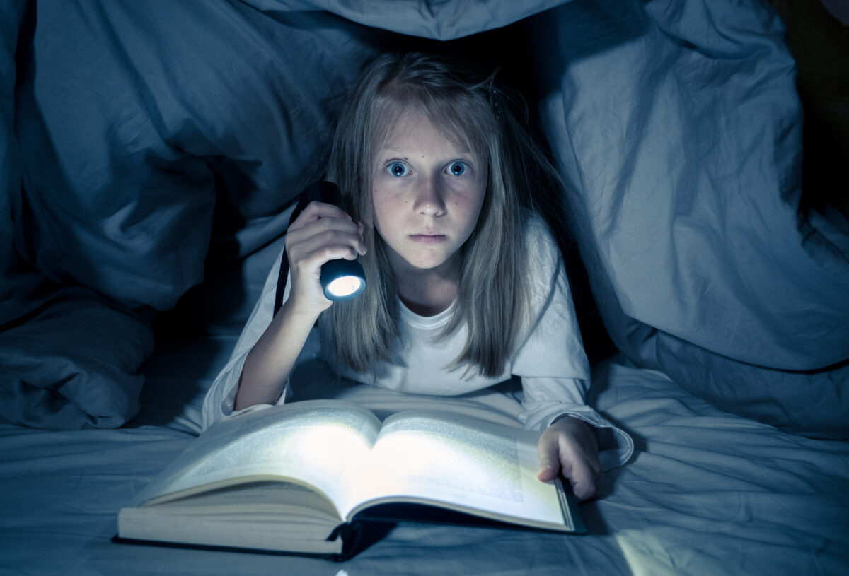 Jente som leser bok under dyna i lyset fra en lommelykt