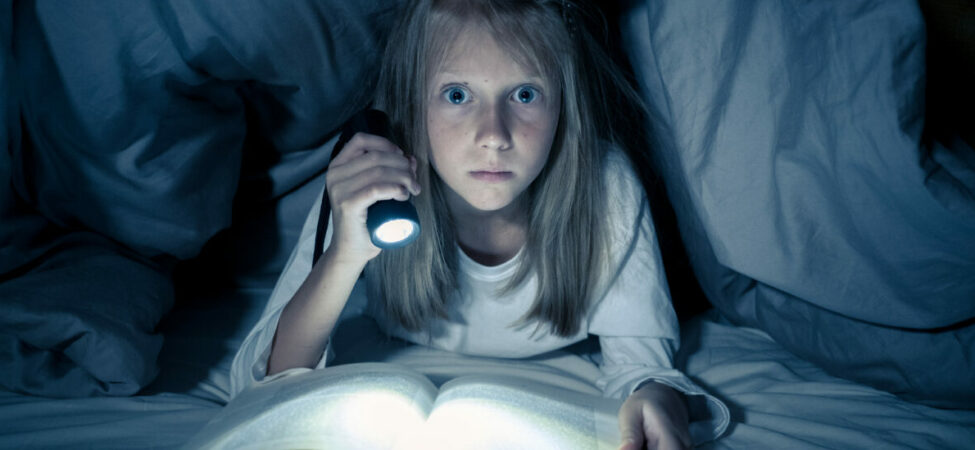 Jente som leser bok under dyna i lyset fra en lommelykt