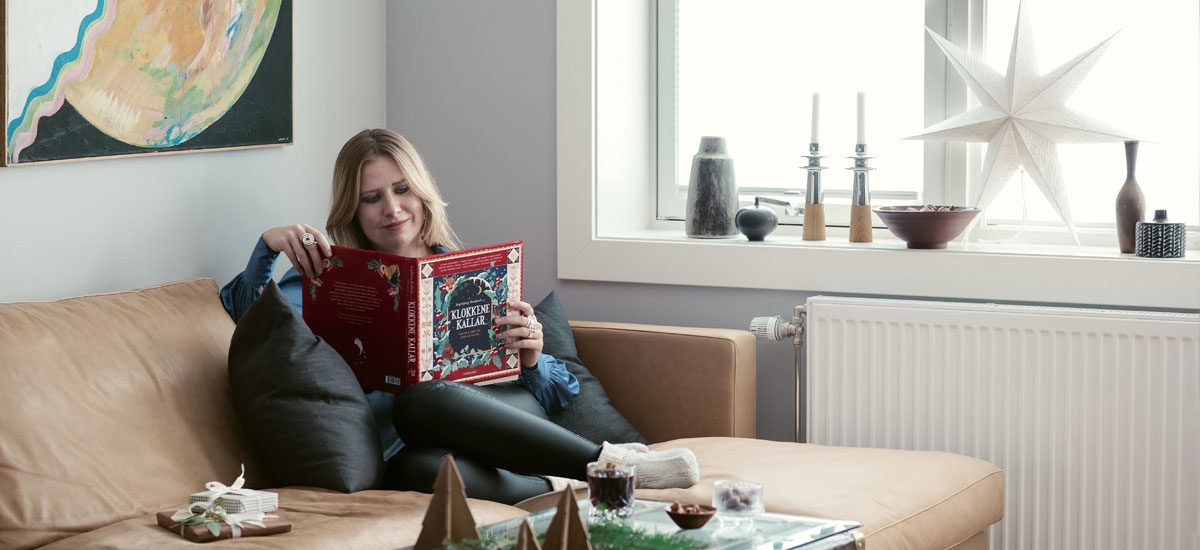 Artist Ingebjørg Bratland sitter i sofaen og leser antologien "Klokkene kallar" som hun har vært redaktør for