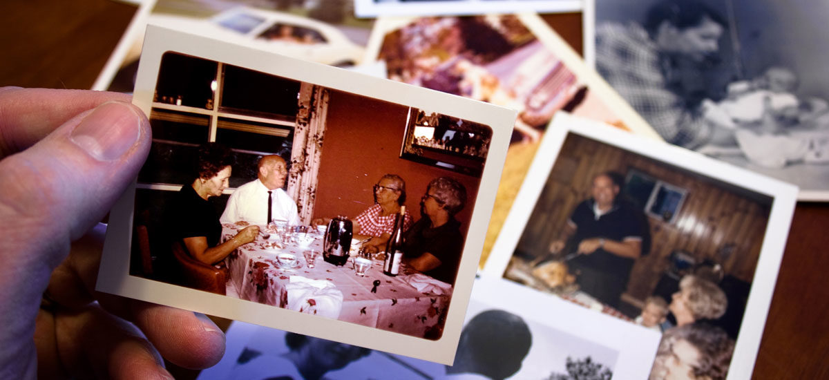 Foto av en hånd som holder et gammelt familiefoto. Andre fotografier på bordet i bakgrunnen.