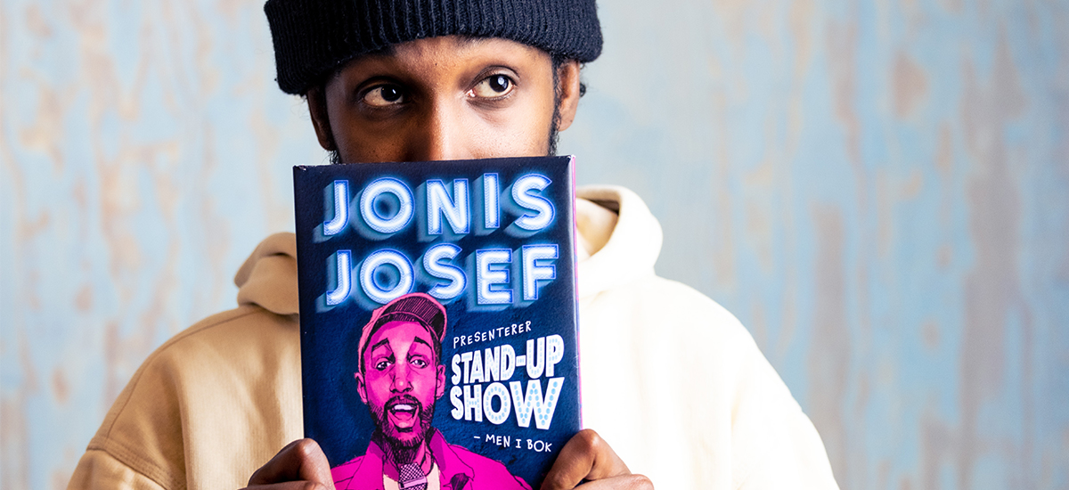 Foto av Jonis Josef med boka «Jonis Josef presenterer stand-up show - men i bok»