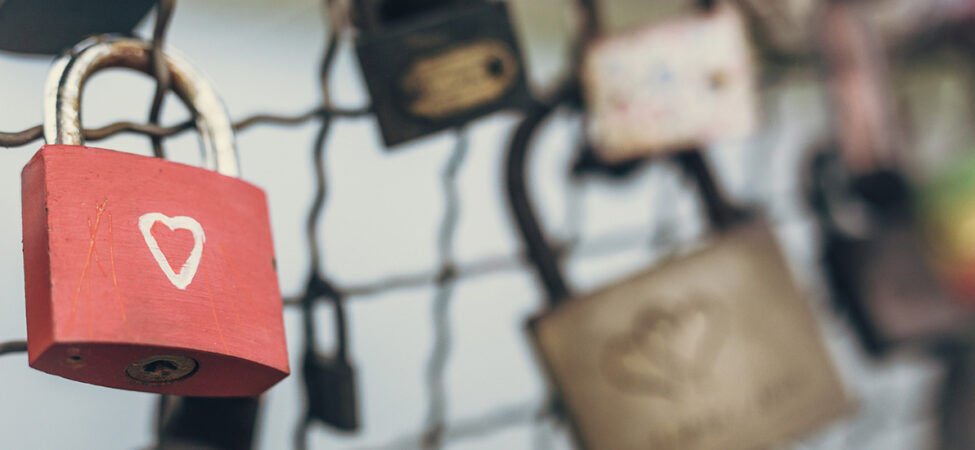 Foto av rød hengelås med hjerte på som henger sammen med mange andre låser på et gjerde
