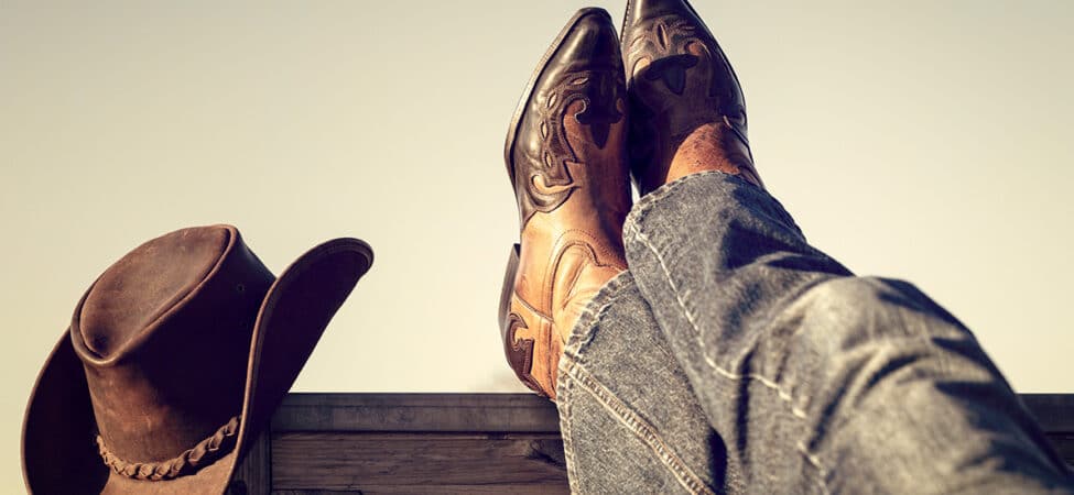 Foto av føttene til en cowboy som hviler på et gjerde. Cowboystøvler og cowboyhatt.